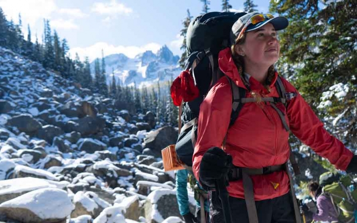 a person carrying a backpack stands on a rocky landscape and smiles contentedly. there are mountains in the background.